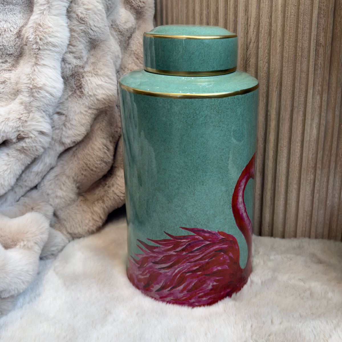 A turquoise porcelain jar with a smooth surface, featuring a stylized flamingo design in red and pink hues. The jar has a wide opening and a matching lid, and is placed on a soft, light-coloured plush surface, with a textured backdrop.