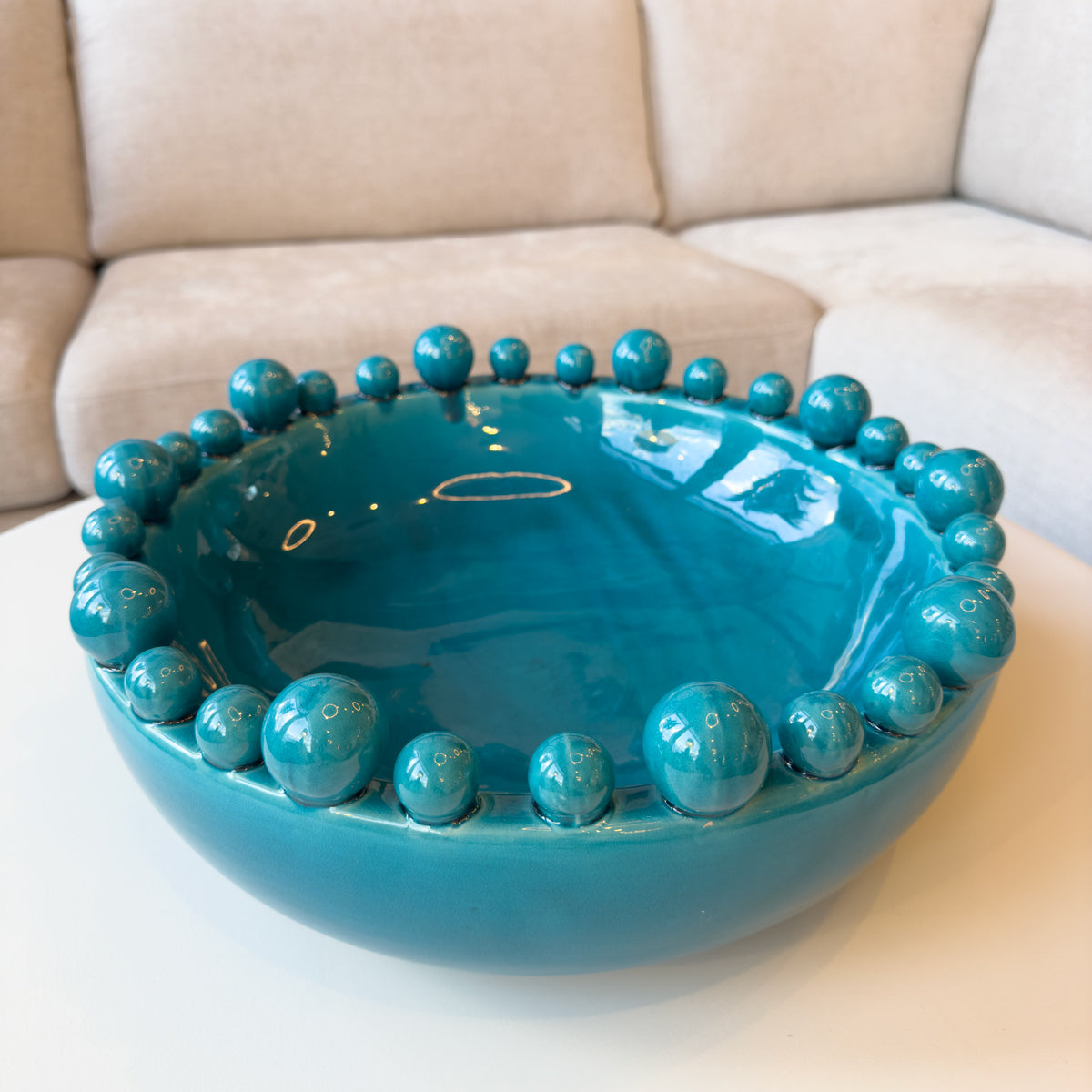 A turquoise green bowl with ball decorations, sitting on a cream coffee table infront of a sofa.