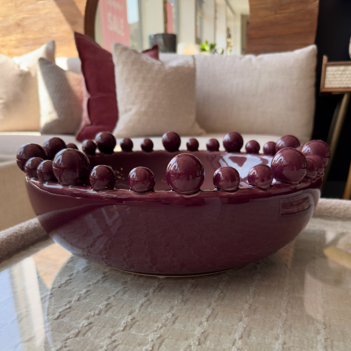 A round, glossy maroon bowl adorned with small round decorative orbs around its rim. The bowl is placed on a light-coloured table, with soft furnishings in the background, including light-coloured cushions and a blurred interior setting.